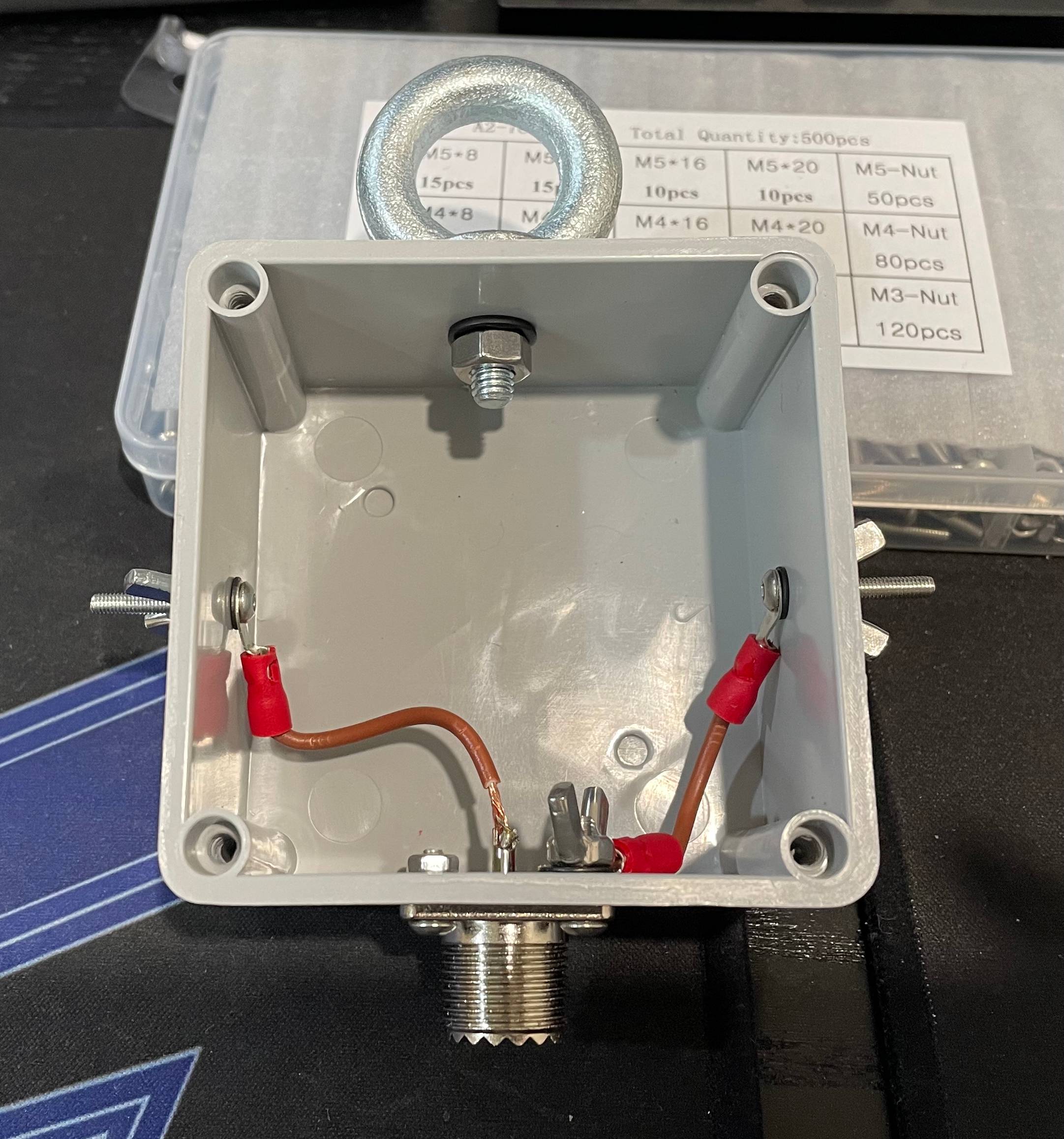 Square plastic antenna box with metal wing nuts drilled into left and right sides, a circular ring eye bolt drilled into the top, and a coaxial connector drilled into the bottom. There are two wires connected to each lug. One wire is badly soldered to the coaxial connector centre pin, and the other is fastened to the coaxial connector shield.