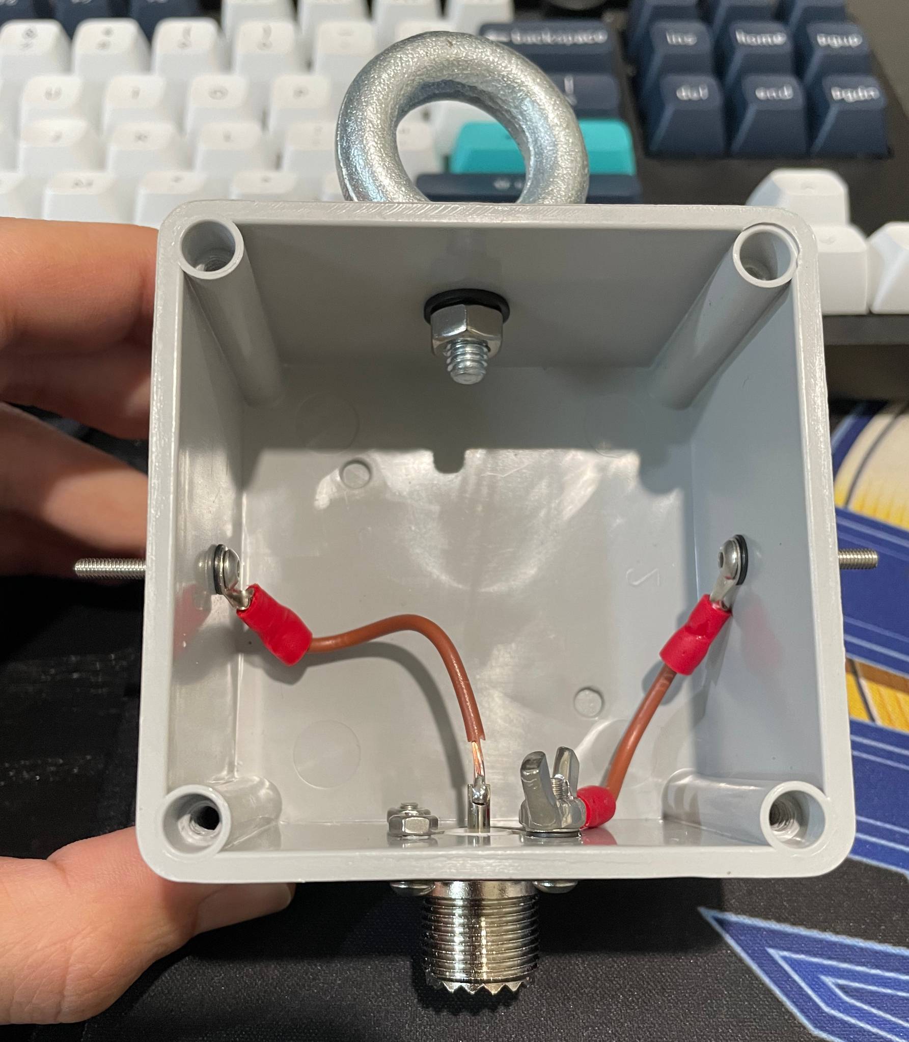 Square plastic antenna box with metal wing nuts drilled into left and right sides, a circular ring eye bolt drilled into the top, and a coaxial connector drilled into the bottom. There are two wires connected to each lug. The photo is highlighting my new solder job on one wire soldered to the coaxial connector centre pin.
